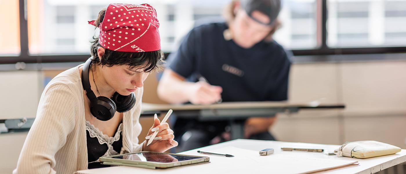 A student in red polka dot bandana and with headphones around their neck uses an iPencil to draw on an iPad.